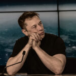Elon Musk in thoughtful pose with hands clasped at a conference or presentation