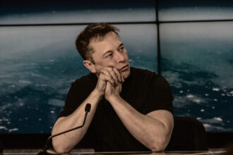 Elon Musk in thoughtful pose with hands clasped at a conference or presentation