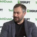 A bearded man grins against a TechCrunch-branded backdrop